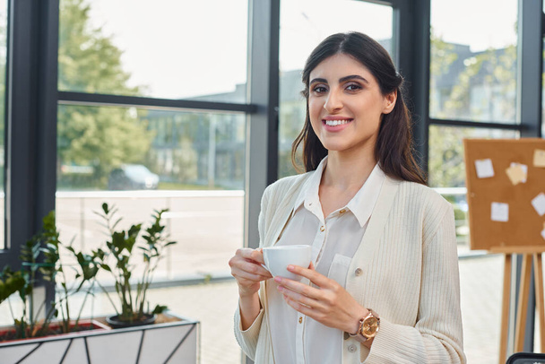 Una mujer de negocios saborea una taza de café contra un telón de fondo de paisaje urbano a través de una gran ventana en una oficina moderna. - Foto, imagen
