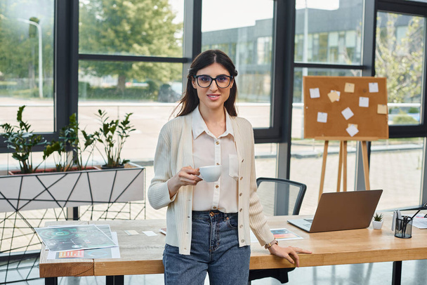 Una mujer de negocios se encuentra en una oficina moderna, enfocada en su computadora portátil en la mesa frente a ella. - Foto, imagen