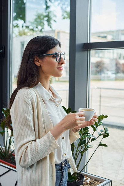 Сучасна бізнес-леді тримає чашку, стоячи перед вікном в офісі, глибоко в думці. - Фото, зображення