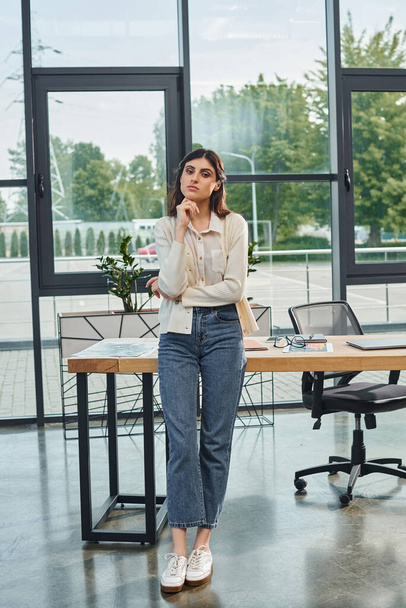 Современная деловая женщина уверенно стоит перед гладким столом в современном офисе. - Фото, изображение