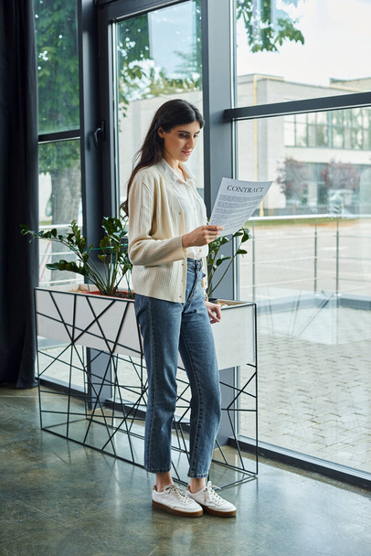 Eine Geschäftsfrau steht vor einem Fenster und hält einen Vertrag in einem modernen Büroraum, tief in Gedanken. - Foto, Bild