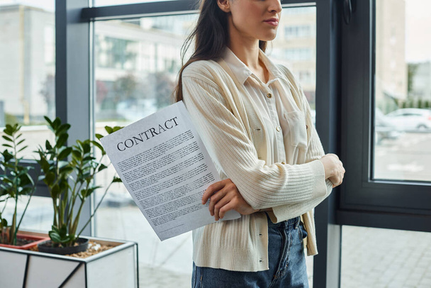 Современная деловая женщина стоит у окна, держа в руках лист бумаги, размышляя о следующих шагах в своей концепции франшизы. - Фото, изображение