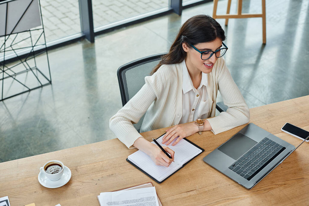 Une femme d'affaires est assise à une table dans un bureau moderne, concentrée sur son ordinateur portable, travaillant sur un concept de franchise. - Photo, image