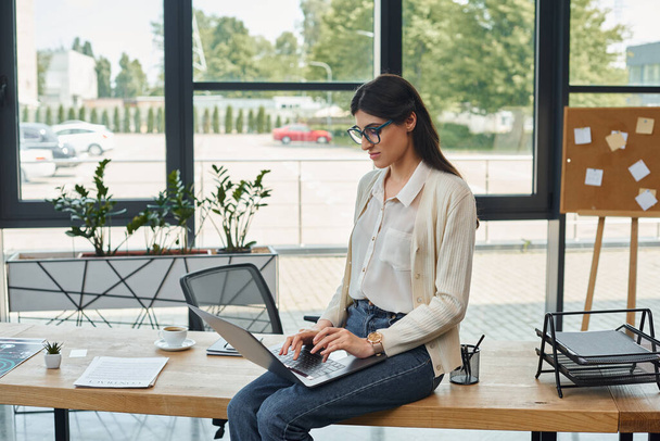 Een zakenvrouw die op een tafel zit met een laptop in een modern kantoor, gericht op franchisewerk. - Foto, afbeelding