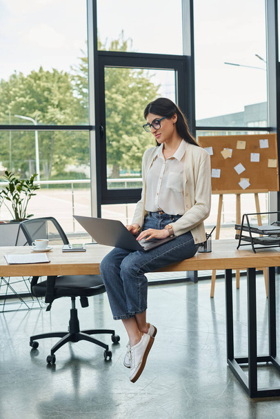 Una donna d'affari in un ufficio moderno siede su un tavolo, lavorando diligentemente sul suo computer portatile. - Foto, immagini