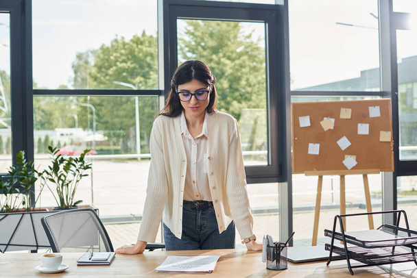 Μια επιχειρηματίας στέκεται με αυτοπεποίθηση μπροστά από ένα κομψό ξύλινο τραπέζι σε ένα σύγχρονο χώρο εργασίας γραφείου. - Φωτογραφία, εικόνα