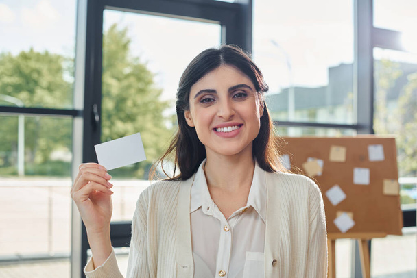 Современная деловая женщина держит визитку перед окном в гладком офисном помещении. - Фото, изображение