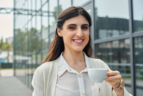 Una donna d'affari elegante in piedi di fronte a un edificio moderno, in possesso di una tazza di caffè. - Foto, immagini