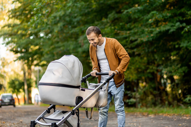 Mutlu bir baba, parkta yürürken bebeğine bakıp bebek arabasını iterken gülümsüyor ve bebeğiyle konuşuyor. - Fotoğraf, Görsel