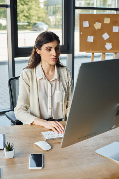 Eine entschlossene Geschäftsfrau sitzt an ihrem Schreibtisch in einem modernen Büro, voll beschäftigt mit ihrem Computer.. - Foto, Bild