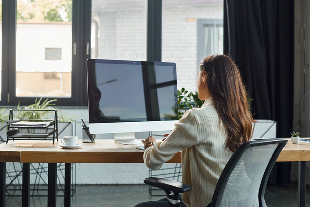 Una donna d'affari immersa nel suo lavoro, seduta alla scrivania in un ufficio moderno, concentrata sullo schermo del computer. - Foto, immagini