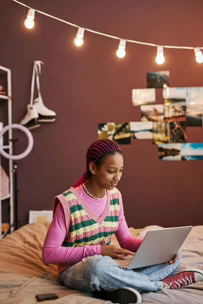 Retrato vertical de adolescente negra sentada na cama com as pernas cruzadas e usando laptop em casa - Foto, Imagem