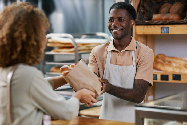 Smiling bakery worker giving package with fresh bread to customer - Photo, Image