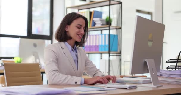 Mujer sonriente se sienta en el escritorio de la oficina y los tipos de informe en la computadora. Joven empresaria con expresión alegre se centra en tareas de cámara lenta - Imágenes, Vídeo