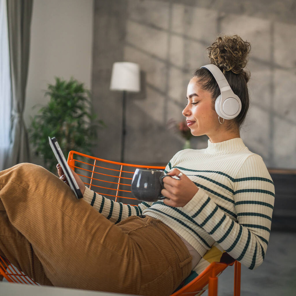 Eine junge Frau sitzt zu Hause und nutzt ein digitales Tablet und Kopfhörer, um Filme oder Serien online zu sehen oder Videoanrufe zu führen. - Foto, Bild