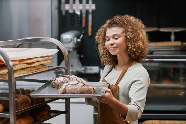 Propietario positivo de una pequeña panadería con cabello rizado mirando bandeja con pan que hizo para vender - Foto, Imagen