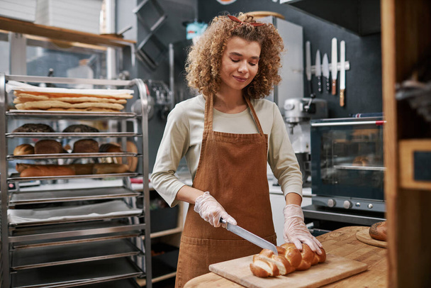 Lächelnder Bäcker mit Handschuhen beim Brotschneiden für Kunden - Foto, Bild