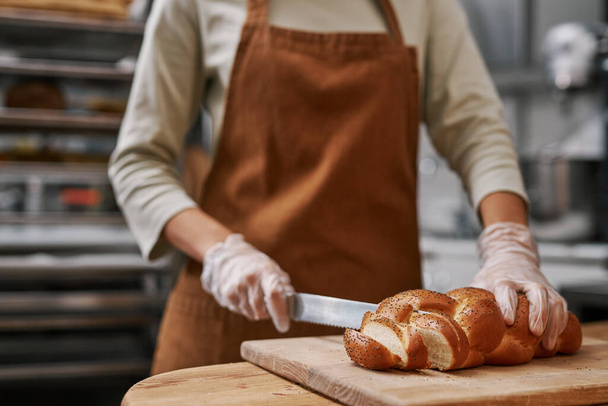Χέρια αρτοποιού που κόβουν πλεγμένο ψωμί με σπόρους παπαρούνας σε λεπτές φέτες - Φωτογραφία, εικόνα