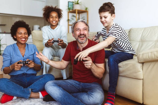 Uśmiechnięta rodzina spędzająca wspólnie czas w domu, siedząc na kanapie w salonie i grając w gry wideo. - Zdjęcie, obraz