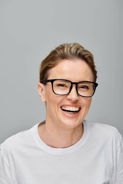 veselá atraktivní žena s blond vlasy s brýlemi šťastně se usmívá na kameru na šedém pozadí - Fotografie, Obrázek