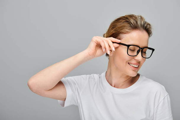 mujer atractiva alegre con el pelo rubio y gafas elegantes posando sobre fondo gris y mirando hacia otro lado - Foto, imagen