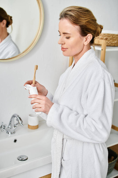 attraktive blonde Frau mit gesammelten Haaren im Bademantel beim Zähneputzen vor dem Spiegel - Foto, Bild