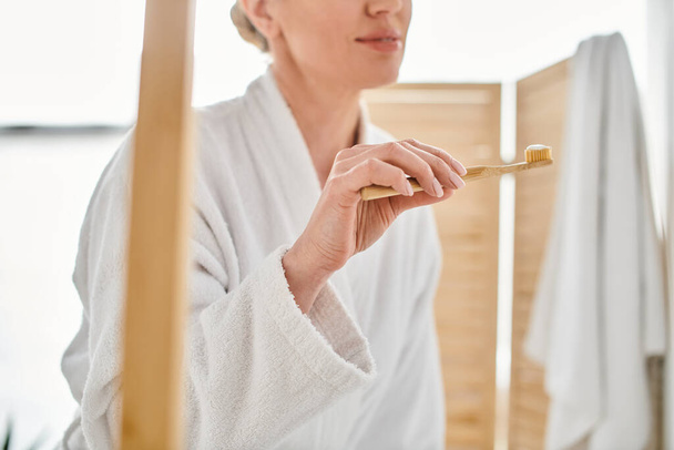 vista recortada de mujer adulta en albornoz acogedor poniendo pasta de dientes en su cepillo cerca del espejo - Foto, imagen