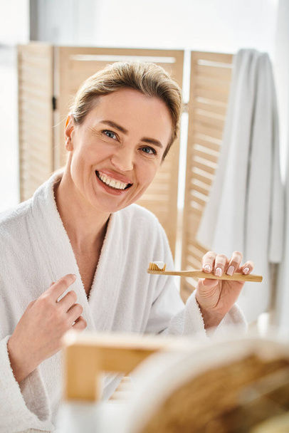 aantrekkelijk vrolijke vrouw in badjas met blond haar op zoek naar camera terwijl poetsen haar tanden - Foto, afbeelding