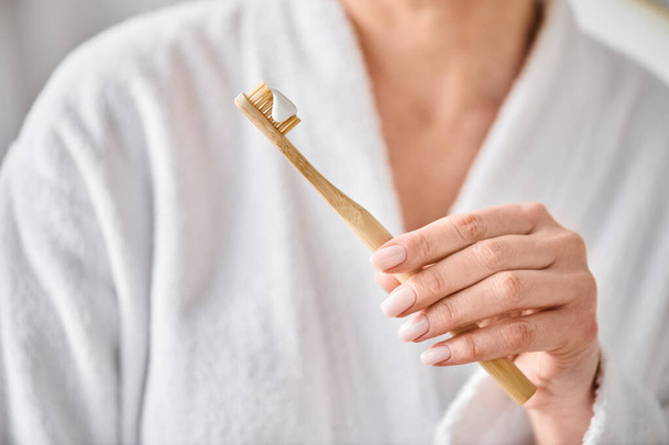 Beyaz bornozlu yetişkin kadın aynanın yanındaki fırçasına diş macunu sürüyor. - Fotoğraf, Görsel