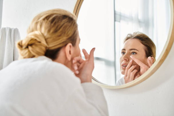 attraktive Frau im weißen bequemen Bademantel mit Kontaktlinsen in der Nähe von Spiegel im Badezimmer - Foto, Bild