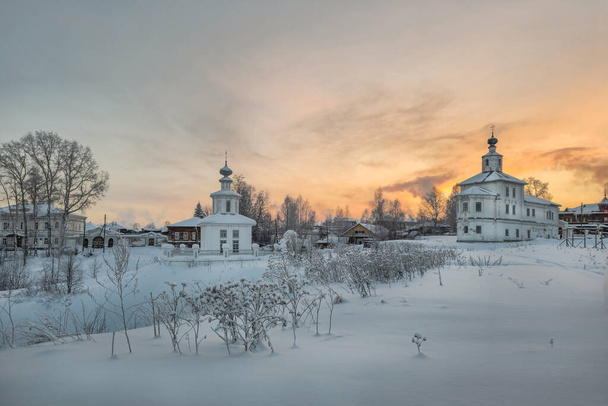 Αρχαία αρχιτεκτονική περιβάλλεται από ψηλά λευκά παρασύρει και πορτοκαλί ουρανό ηλιοβασίλεμα. Ιστορικό κέντρο της αρχαίας πόλης Cherdyn, Ρωσία στα μέσα του χειμώνα. Παρεκκλήσι Spasskaya Cherdyn.  - Φωτογραφία, εικόνα