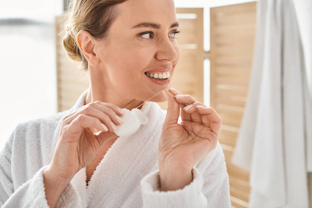 χαρούμενη ελκυστική γυναίκα με λευκό μπουρνούζι που καθαρίζει τα δόντια της με οδοντικό νήμα στο μπάνιο - Φωτογραφία, εικόνα