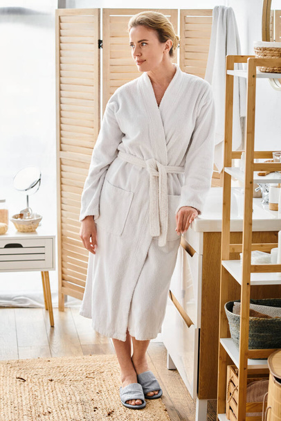 приваблива весела жінка з білявим волоссям у білому халаті позує у своїй ванній кімнаті і дивиться - Фото, зображення