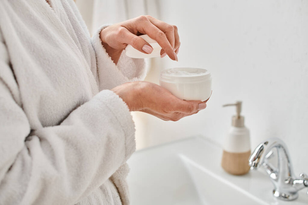 Ausgeschnittene Ansicht einer erwachsenen Frau im bequemen Bademantel mit Gesichtscreme in den Händen im Badezimmer - Foto, Bild