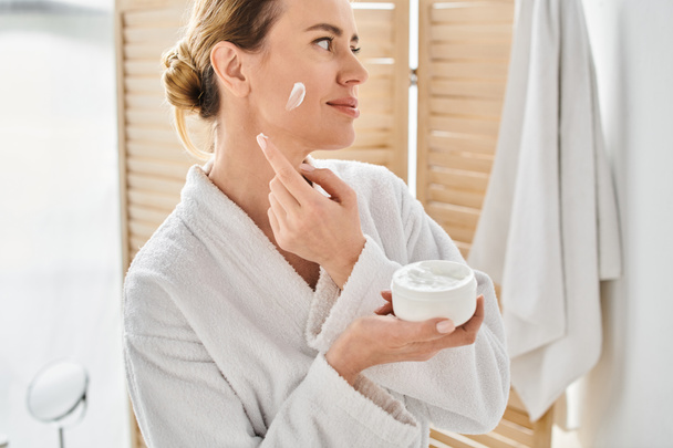 attraente donna bionda in comodo accappatoio bianco applicare la crema per il viso mentre in bagno a casa - Foto, immagini