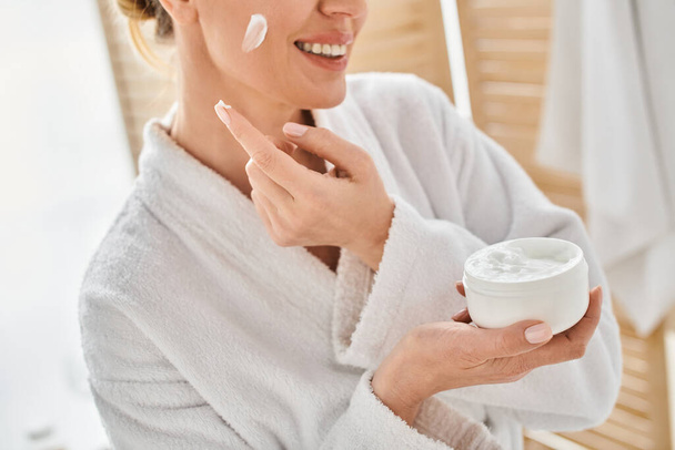ostříhaný pohled na veselou ženu v pohodlném županu použití její obličej krém v koupelně u ní doma - Fotografie, Obrázek