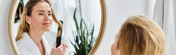 mulher loira atraente alegre em roupão de banho acolhedor usando ferro liso em seu cabelo enquanto no banheiro - Foto, Imagem