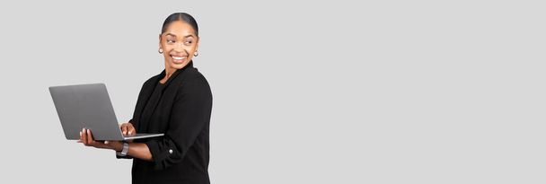 Una donna d'affari afro-americana allegra tiene un computer portatile con una mano mentre sorride e guarda di lato, ritraendo un senso di facilità e competenza, isolato su sfondo grigio studio - Foto, immagini
