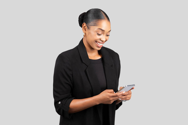 Зміст молодої професійної жінки в офіційному вбранні захоплюється її смартфоном, можливо, керує діловими комунікаціями з посмішкою, ізольовані на сірому студійному фоні - Фото, зображення