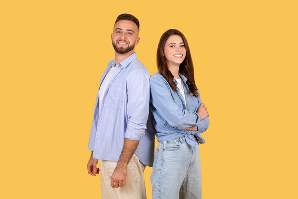 Souriant jeune homme et femme caucasien en tenue décontractée se tenir dos à dos, exsudant confiance et bonheur sur fond jaune vibrant - Photo, image