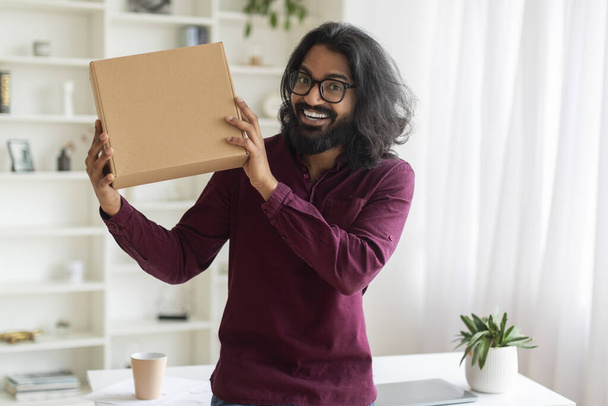 Cheerful indian man holding cardboard box while standing near desk at home, happy eastern male exuding excitement for an unboxing video, enjoying fast delivery services, copy space - Photo, Image