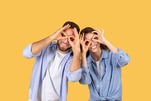 Riendo hombre y mujer juguetonamente haciendo formas binoculares con sus manos alrededor de sus ojos, compartiendo momento divertido en el fondo amarillo brillante - Foto, imagen