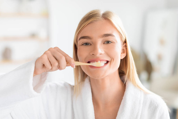 若い金髪の女性は,現代のバスルームで朝のルーチンの一部として口腔衛生に焦点を当てて歯を磨いています. 彼女の歯のクレンジングのための歯ブラシの世話を持つ女性の肖像画ショット - 写真・画像
