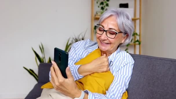 Donna anziana felice utilizzando il telefono cellulare mentre si rilassa a casa. Femmina dai capelli grigi degli anni '60 che sorride guardando lo schermo dello smartphone. Concetto di tecnologia e anziani. - Filmati, video