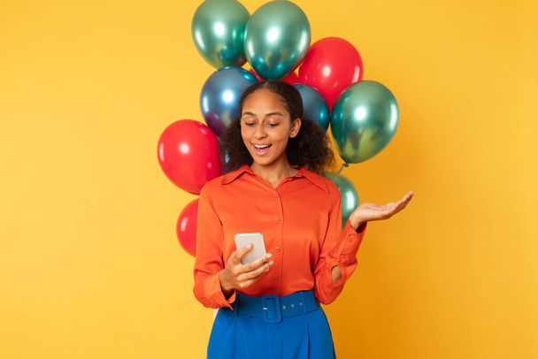 Joyeux jeune fille noire tenant smartphone message de lecture, posant avec un tas de ballons colorés lumineux, navigation offres festives sur son anniversaire sur fond de studio jaune - Photo, image