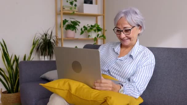 Взрослая женщина 60-х годов использует ноутбук, сидя дома на диване. Вышедшая на пенсию седовласая женщина в очках, печатающая на ноутбуке, занимающаяся серфингом в сети.  - Кадры, видео
