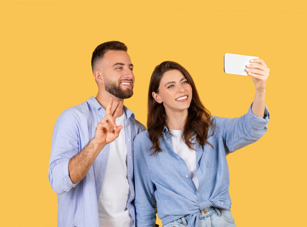 Улыбающиеся мужчина и женщина делают селфи вместе, мужчина мигает знак мира, оба наслаждаются веселым моментом, запечатленным на смартфоне на ярко-желтом фоне - Фото, изображение
