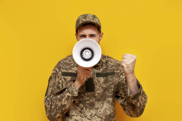 Oekraïense soldaat in camouflage uniform kondigt informatie in een luidspreker op een gele geïsoleerde achtergrond, een mannelijke militaire man schreeuwt in een megafoon - Foto, afbeelding