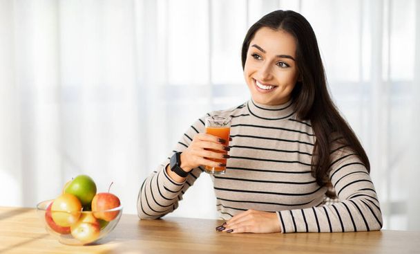Jovem alegre mulher do Oriente Médio desfrutando de um copo fresco de suco, com uma tigela de frutas coloridas na mesa, promovendo um estilo de vida saudável e nutrição na cozinha doméstica - Foto, Imagem