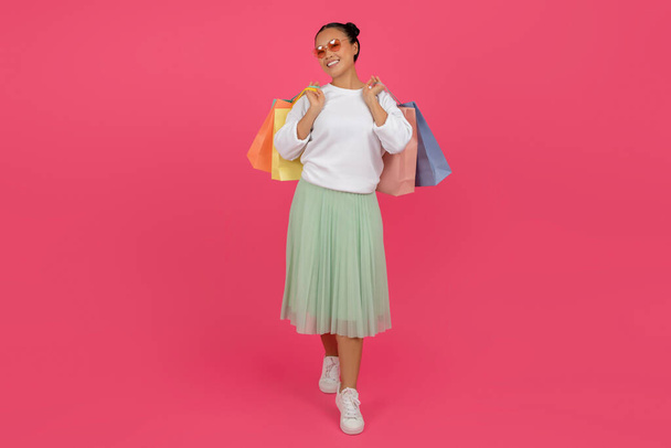 Gelukkig aziatische vrouw wandelen met helder winkelen tassen in handen op roze achtergrond, vrolijke glimlachende vrouw dragen aankopen en kijken naar camera, genieten van zwarte vrijdag verkoop, volledige lengte, kopiëren ruimte - Foto, afbeelding